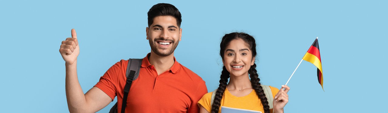 Ein junger Mann und eine junge Frau stehen nebeneinander vor einem hellblauen Hintergrund. Beide lächeln. Die Frau hält zusätzlich eine kleine deutsche Flagge.