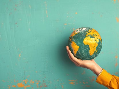 Eine Hand hält eine kleine Weltkugel vor einem türkisfarbenen, leicht strukturierten Hintergrund. Auf der Kugel sind Afrika, Europa und Teile Asiens zu sehen.