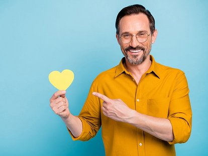 Mann mittleren Alters mit Brille und Bart hält ein gelbes Papierherz in der Hand und zeigt mit dem Finger darauf. Er trägt ein senfgelbes Hemd und lächelt. Hintergrund ist einfarbig hellblau.