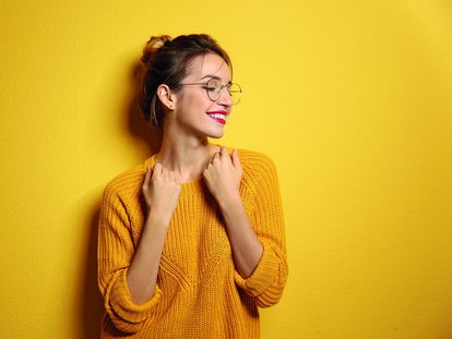 Junge Frau mit Brille und hochgesteckten Haaren trägt einen gelben Strickpullover und lächelt verträumt zur Seite. Sie steht vor einer gelben Wand.
