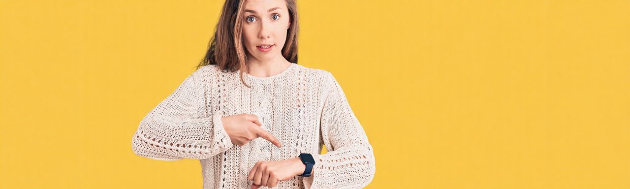 Junge Frau mit langen Haaren und hellem Strickpullover zeigt mit dem Finger auf ihre Smartwatch am Handgelenk. Sie schaut überrascht oder fragend in die Kamera. Hintergrund ist einfarbig gelb.
